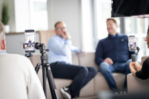 Ein Behind-the-scenes Foto, das eine Interviewaufnahme mit einem Smartphone auf einem Stativ zeigt. Im unscharfen Hintergrund sitzen Steven Klatt und Gerd Schmees auf einem Sofa.