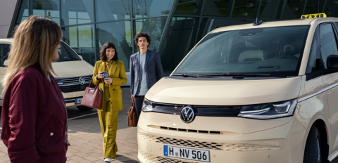 Ein hellbeiger Volkswagen Multivan als Taxi steht vor einem modernen Glasgebäude, während zwei Fahrgäste auf das Fahrzeug zugehen.