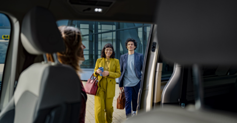 Blick aus dem Innenraum eines Volkswagen Taxis auf zwei Fahrgäste, die lächelnd auf das Fahrzeug zugehen.