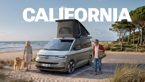 VW California Campervan mit aufgestelltem Schlafdach an einem Sandstrand, daneben ein Paar mit Hund. Schriftzug „CALIFORNIA“.