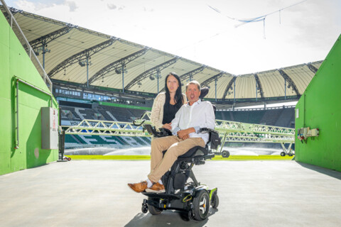 Philipp Hanf in seinem Elektrorollstuhl und eine junge Frau, die lächelnd hinter ihm steht, im Eingangsbereich eines Fußballstadions.